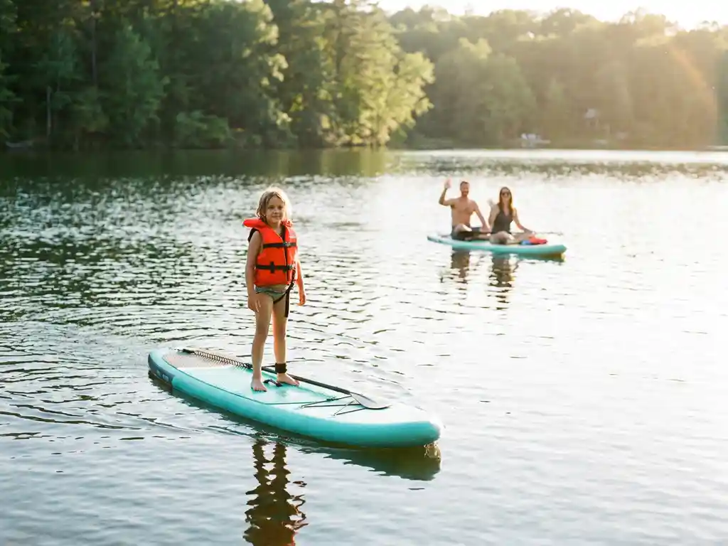 Kind van 8 jaar balanceert op turquoise SUP-board op kalm meer, oranje zwemvest, ouders op achtergrond, gouden zonlicht.