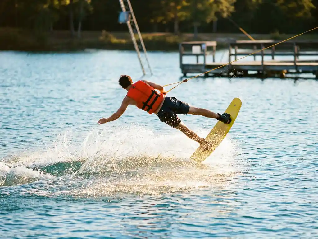 Wakeboarder valt in helder blauw meerwater, oranje zwemvest en geel board zichtbaar, gouden zonlicht weerkaatst op wateroppervlak