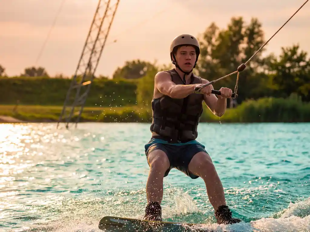 Wakeboard beginner met gespannen greep op kabeltouw, licht gebogen knieën, glijdend over fonkelend turquoise water bij gouden uurtje