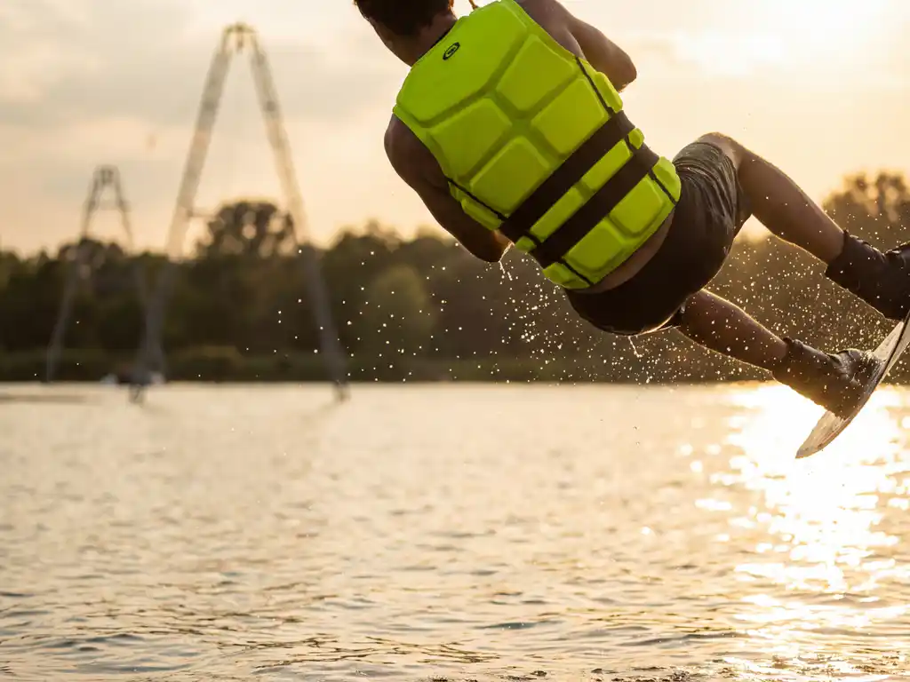 Wakeboarder in felgele impact vest springt boven glinsterend meer met waterdruppels in de lucht