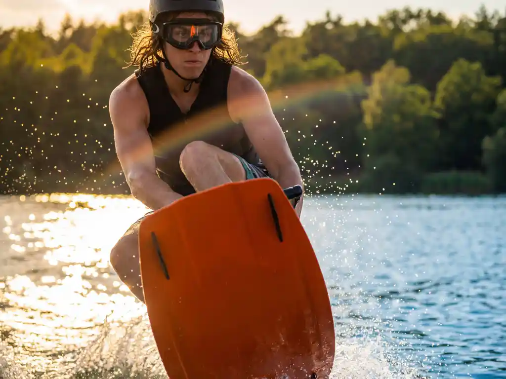Wakeboarder springt boven fonkelend meer met zwarte sportbril, oranje wakeboard zichtbaar, Belgisch bos op achtergrond